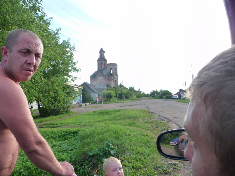 Pavel grimacing at Evgeny explaining the confluence concept to him. View of hamlet, dilapidated cathedral and our white jeep Niva in the background.