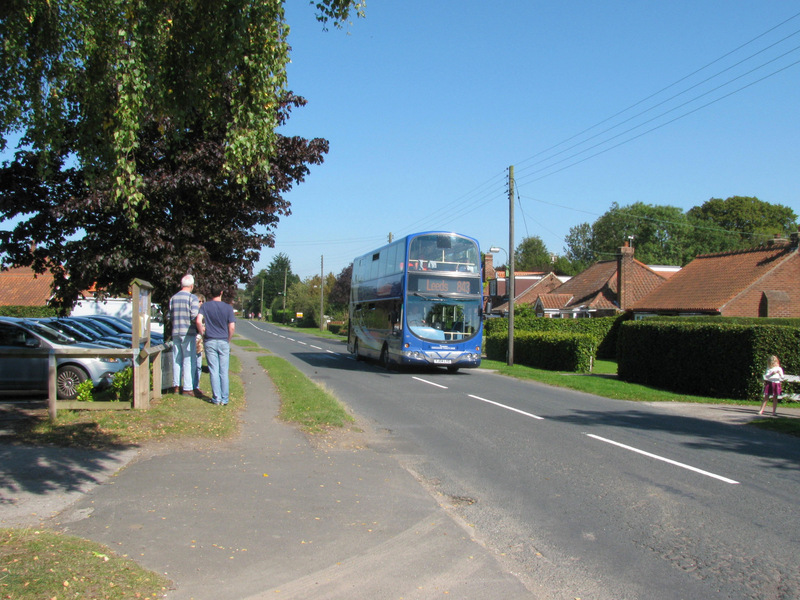 A double decker bus drives along Stockton-on-the-Forest's one street.  People are gathering to watch the "Tour of Britain" bike racers.
