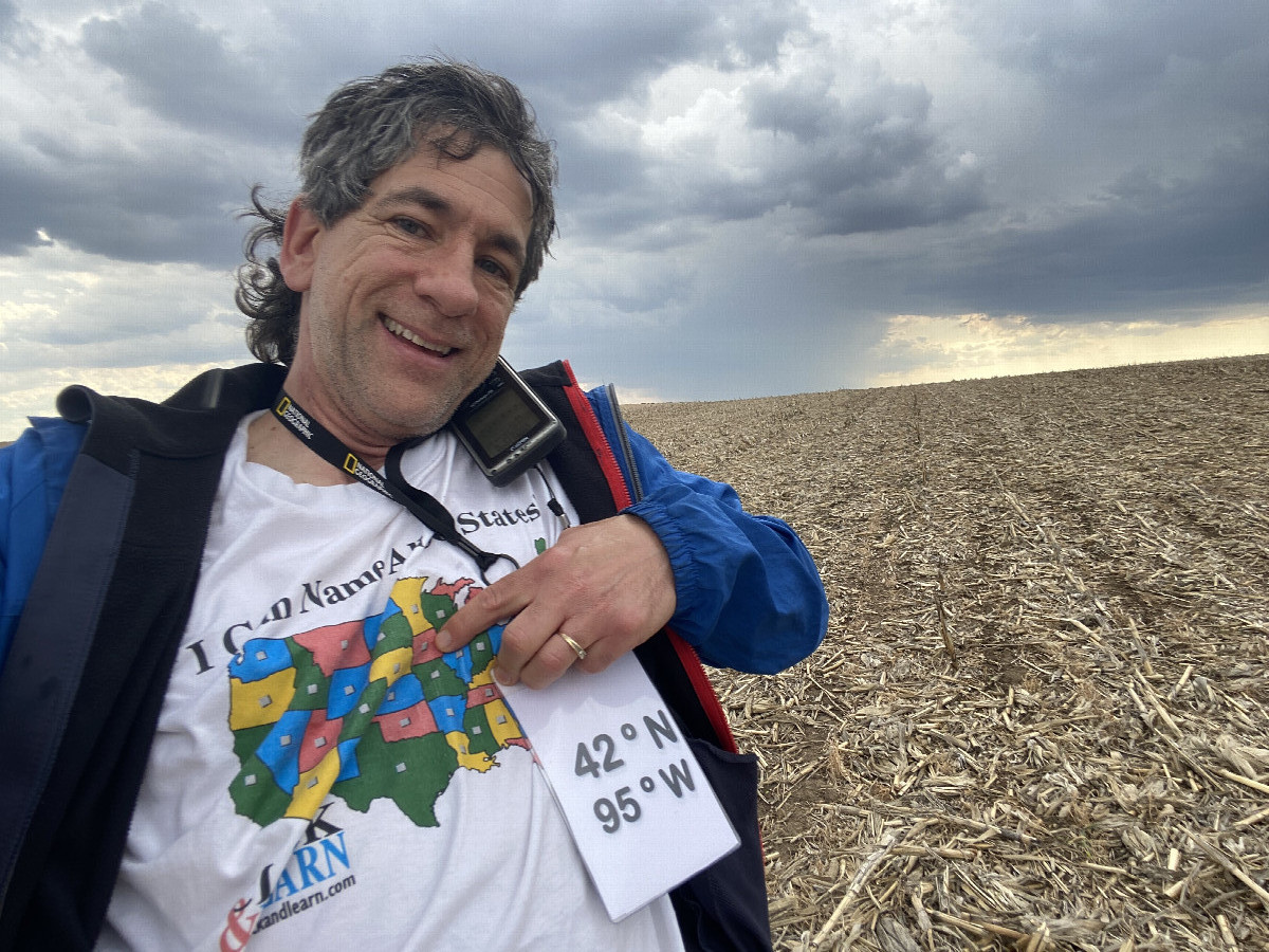 Joseph Kerski at the confluence point, pointing at Iowa on map shirt. 